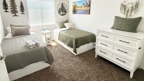 Un dormitorio con cama, cómoda, mesita de noche, ventana con persianas, ventilador de techo y decoraciones en la pared, incluyendo siluetas de árboles y un cuadro de paisaje.