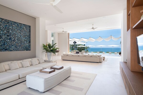 A modern living room with a large sofa, a coffee table, and a TV. There is a large painting on the wall and a potted plant. The room has a view of the ocean and outdoor seating. - Estate Buho in Punta Mita (Punta de Mita)