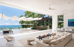 A modern living room with a large sofa, a coffee table with books, a pool, and a view of the ocean. - Estate Buho in Punta Mita (Punta de Mita)