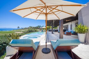 Ein Poolbereich mit Liegestühlen, einem großen Sonnenschirm und Blick aufs Meer.