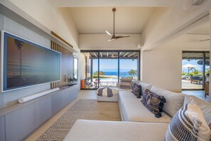 Ein modernes Wohnzimmer mit einem großen Fernseher, einem Deckenventilator und Blick aufs Meer.