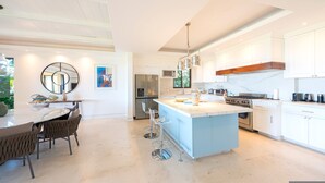 A modern kitchen with white cabinets, a central island, stainless steel appliances, and a dining area with a round mirror and artwork. - Condo Zazil in Punta Mita (Punta de Mita)