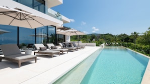 Un área moderna junto a la piscina con sillones de descanso y sombrillas, una piscina cristalina y vistas a zonas verdes y colinas.