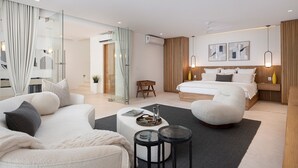 Una habitación de hotel moderna con una cama grande, una zona de estar con sofá y sillón, una mesita y un área de ducha con mampara de vidrio.