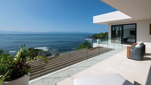 Un balcón moderno con vistas al océano, una puerta de vidrio que da al interior y un área de estar con cojines.