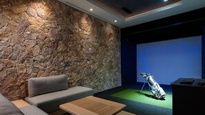 Une pièce avec un mur en pierre, un canapé gris, une table basse en bois et un écran qui affiche un parcours de golf.