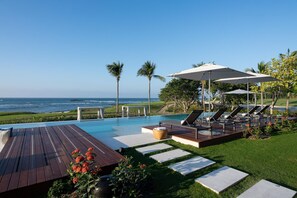Uma área à beira da piscina com espreguiçadeiras, guarda-sóis e vista para o oceano.