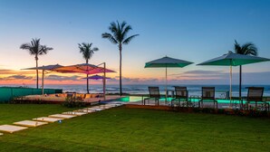 Un espace aménagé au bord de la piscine, avec des chaises longues et des parasols, des palmiers, et une vue sur l’océan au coucher du soleil.
