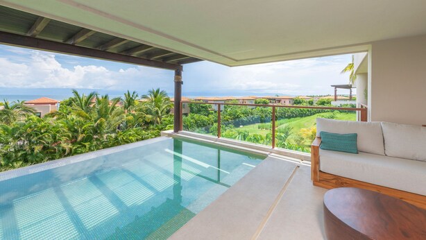 Ein moderner Infinity-Pool mit freiem Blick auf das Meer und Palmen, eine weiße Couch mit einem türkisfarbenen Kissen und ein Holztisch.