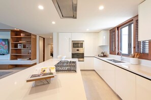 A modern kitchen with white cabinetry, a central island with a stove, and a window with wooden shutters. - Condo Nova in Punta Mita (Punta de Mita)