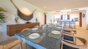 Een eethoek met een lange tafel gedekt voor zes personen, een keuken op de achtergrond en een hangmat aan de muur.