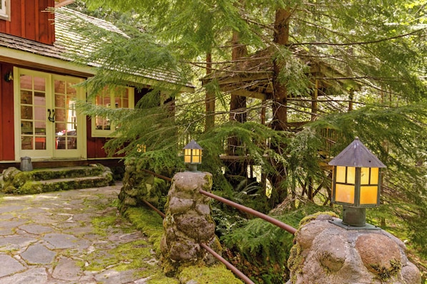 A stone pathway leading to a wooden building with a green door, surrounded by lush greenery and a stone lantern post.
