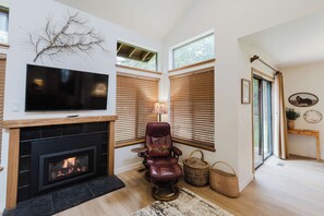 A living room with a fireplace, a television, a leather chair, and large windows. - 3BR Townhouse Near SHARC & Village - Dog Friendly (Sunriver)