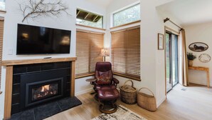 Een woonkamer met een open haard, een televisie, een leren fauteuil en grote ramen.