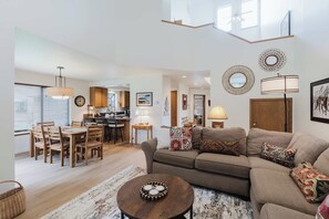 A spacious living area with a sofa, coffee table, dining table, and chairs. There is a kitchen in the background, a dining area with a chandelier, and a large mirror on the wall. - 3BR Townhouse Near SHARC & Village - Dog Friendly (Sunriver)