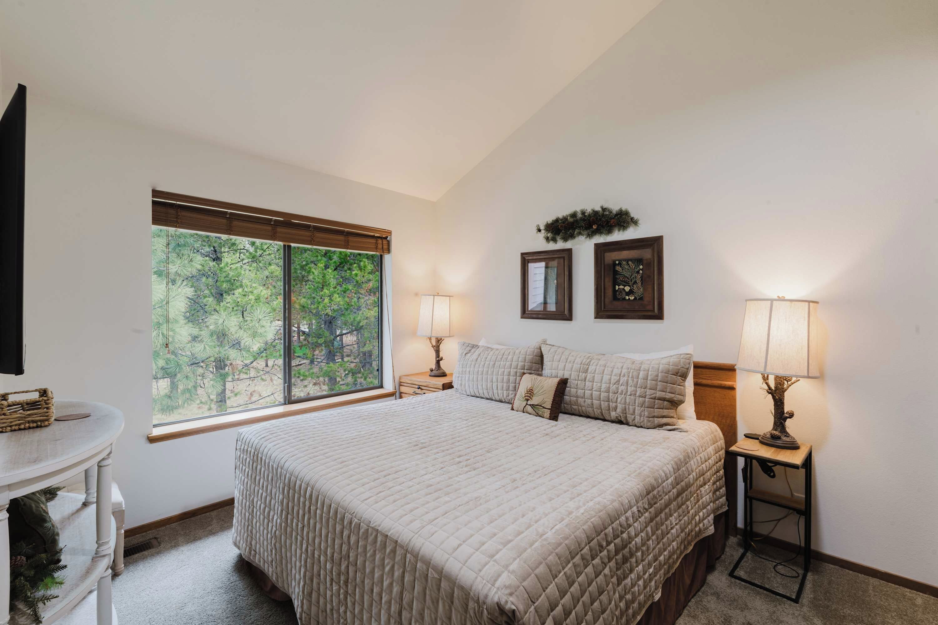 A bedroom with a large bed, two bedside tables with lamps, a window with a view of trees, and a small table with a basket.