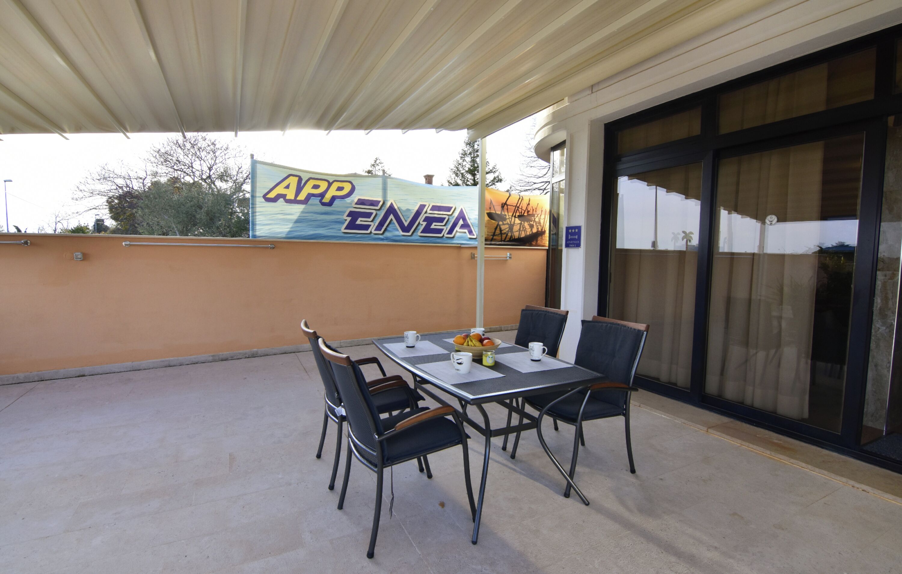 An outdoor patio with a table and chairs, a sign reading 'APP ENEA', and a building entrance with glass doors.