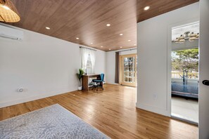 A spacious room with wooden flooring, a desk with a chair, a potted plant, and a large window with a view of the outdoors.