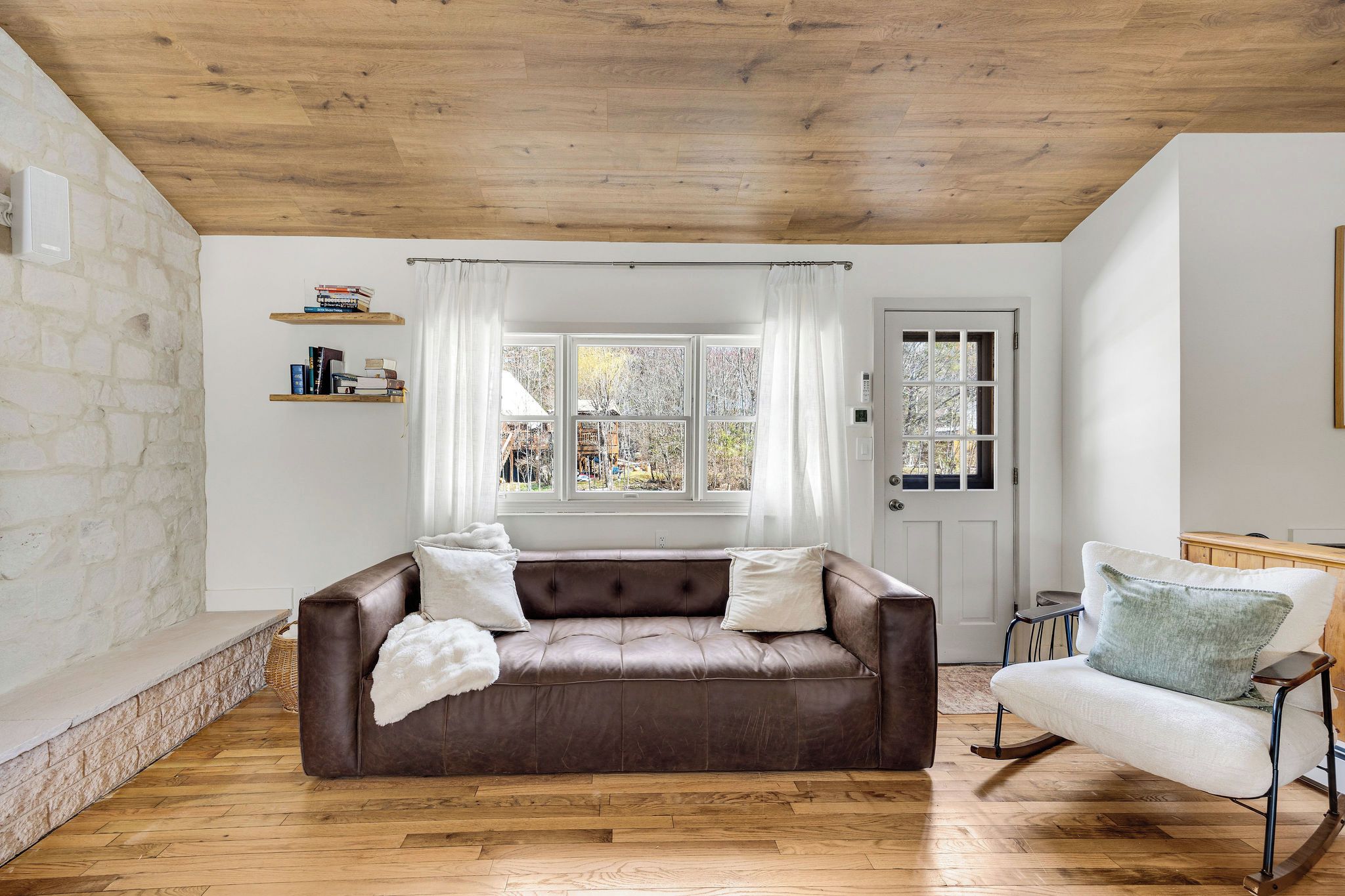 A living room with a brown leather sofa, a white armchair, a wooden floor, a stone wall, and a window with curtains.