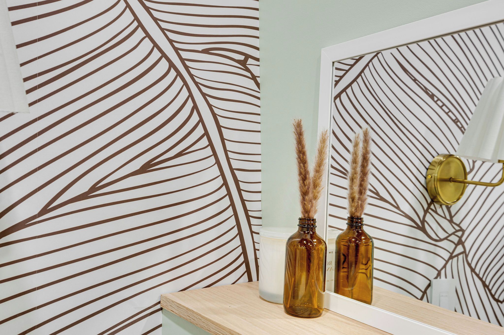 A mirror reflecting a wall with a leaf pattern, a candle, and two bottles with dried plants.