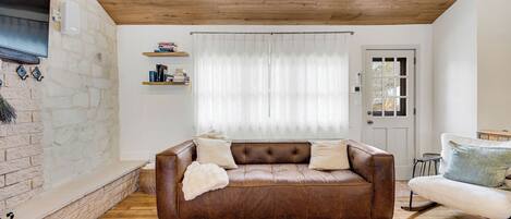 Ein Wohnzimmer mit einem braunen Ledersofa, einer weißen Tür und einem Fenster mit durchscheinenden Vorhängen.