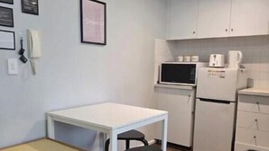 Een kleine keuken met een witte tafel, een bankje, een magnetron, een koelkast en een prullenbak.