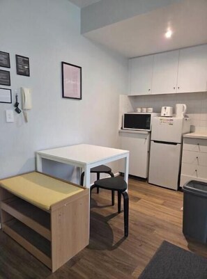 A compact kitchen with a white table, a bench, a microwave, a refrigerator, and a trash bin.