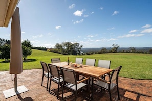 An outdoor patio with a wooden table and chairs, a bottle of wine, and a view of a grassy field and trees. - Wingana Homestead - Scenic Views, Bundanoon (Wingecarribee)