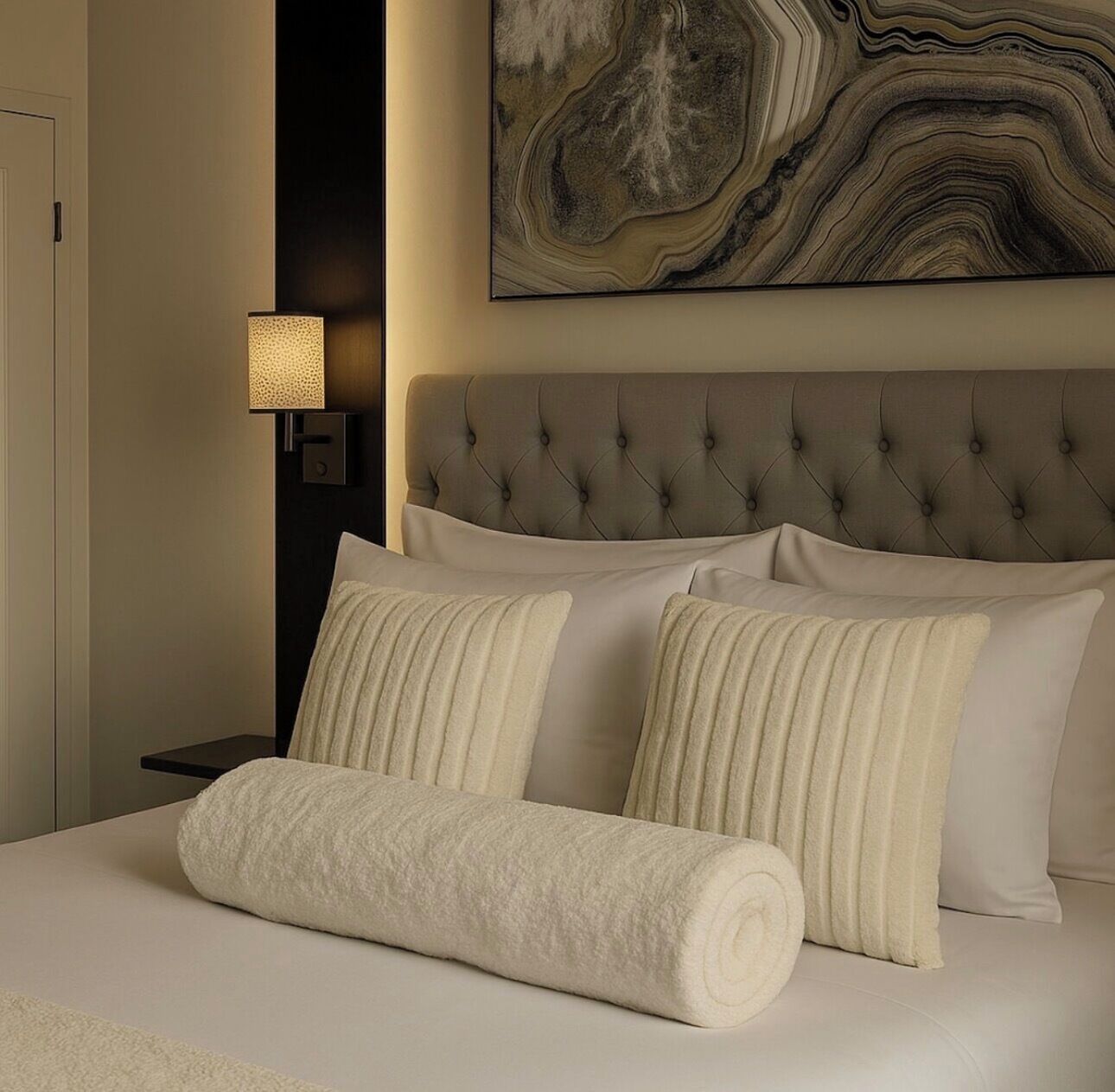 A neatly made bed with a rolled towel, a tufted headboard, and a wall-mounted lamp.