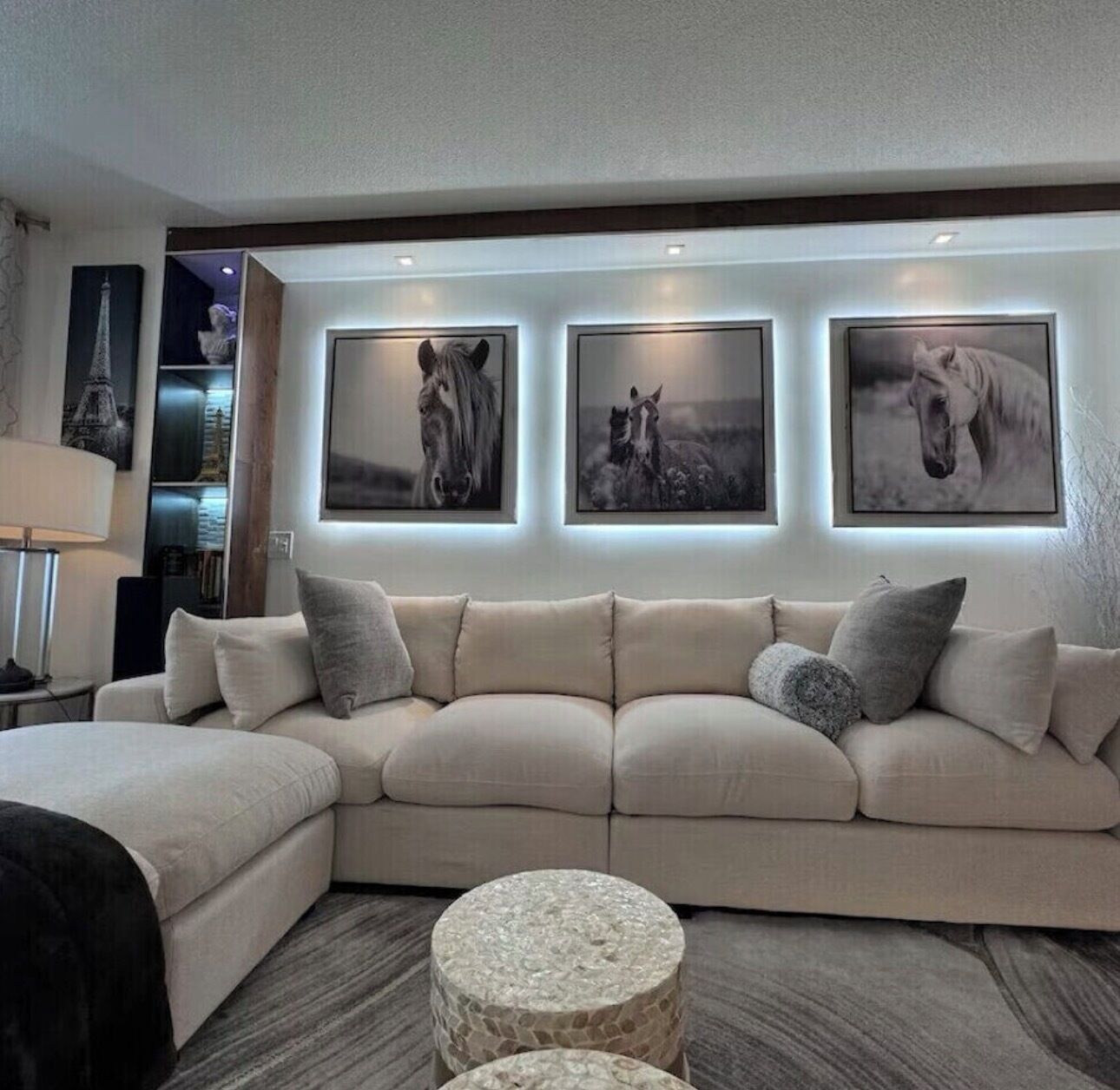 Een moderne woonkamer met een beige hoekbank, drie ingelijste paardenportretten, een stenen salontafel en een lamp.
