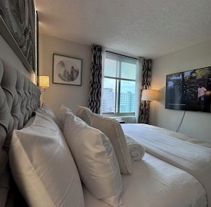 Habitación de hotel con cama, televisor, ventana con cortinas e imagen en un marco en la pared.