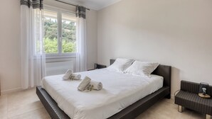 Une chambre à coucher avec un grand lit, du linge de lit blanc, une table de chevet en osier et une vue sur la verdure.