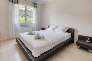 Une chambre à coucher avec un grand lit, du linge de lit blanc, une table de chevet en osier et une vue sur la verdure.