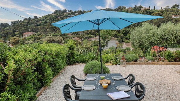 Une table installée à l’extérieur, protégée par un parasol bleu, entourée de gravier et de verdure, et offrant une vue sur une colline parsemée de maisons.