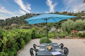 A table set outdoors with a blue umbrella, surrounded by gravel and greenery, overlooking a hillside with houses.