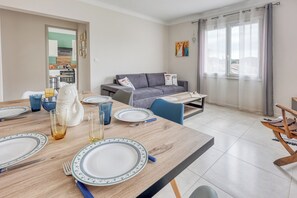 A dining area with a wooden table set for four, a grey sofa, and a view through a window with curtains.