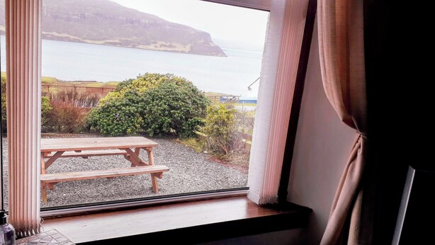 A window with a view of a wooden picnic table, gravel ground, and a bush.