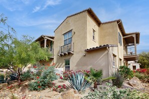 A two-story building with balconies, surrounded by a landscaped garden with various plants and flowers. - Casa Gregario - Beautiful Condo in Spanish Walk Neighborhood (Palm Desert)