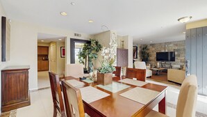 Ein Essbereich mit Holztisch und Stühlen, im Hintergrund eine Küche und ein Wohnzimmer mit Fernseher und Steinwand.