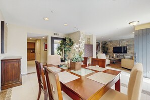 A dining area with a wooden table and chairs, a kitchen in the background, and a living room with a TV and stone wall. - Casa Gregario - Beautiful Condo in Spanish Walk Neighborhood (Palm Desert)