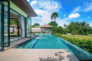 A modern swimming pool with glass walls, a covered seating area, and lush greenery. - Banyan Tree Grand Residences by Banyan Living (Choeng Thale)