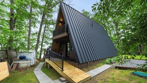 A modern, triangular-shaped cabin with a wooden deck and a small balcony, surrounded by lush greenery. - Lakefront A-Frame | Hot Tub, Dock & Views | Seneca Lake (Penn Yan)