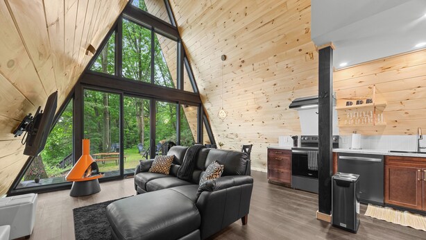 A modern interior with a large living area, a kitchen, and large windows offering a view of the outdoors. - Lakefront A-Frame | Hot Tub, Dock & Views | Seneca Lake (Penn Yan)