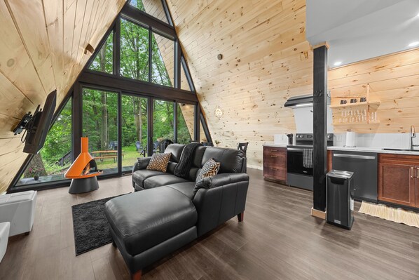 A modern interior with a black leather sofa, wooden ceiling, and a kitchen area. - Lakefront A-Frame | Hot Tub, Dock & Views | Seneca Lake (Penn Yan)