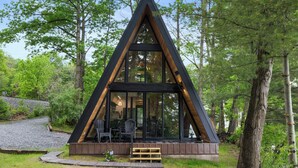 A modern A-frame cabin with large windows, a deck, and a surrounding forest. - Lakefront A-Frame | Hot Tub, Dock & Views | Seneca Lake (Penn Yan)