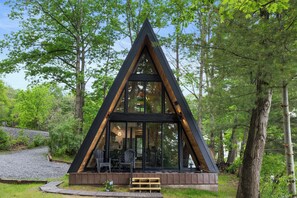 A triangular wooden cabin with large windows, a wooden deck, and a view of the surrounding forest. - Lakefront A-Frame | Hot Tub, Dock & Views | Seneca Lake (Penn Yan)