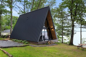 A modern cabin with a sloped roof, a wooden deck, and a view of the lake. - Lakefront A-Frame | Hot Tub, Dock & Views | Seneca Lake (Penn Yan)