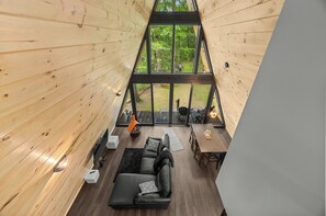 A wooden cabin interior with a large glass window, a black leather sofa, a dining table, and a small kitchen area. - Lakefront A-Frame | Hot Tub, Dock & Views | Seneca Lake (Penn Yan)