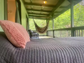 Una cama con cubierta gris texturizada y almohada rosa, una hamaca y vistas a la vegetación por una ventana.