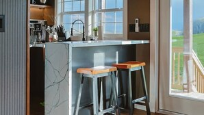 Una cocina moderna con una isla de mármol, taburetes de bar y ventanas amplias.